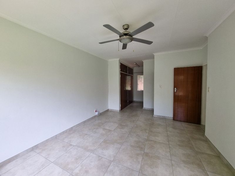 An empty living area featuring neutral tiled flooring, a ceiling fan, and built-in storage cupboards, offering a clean and versatile space.