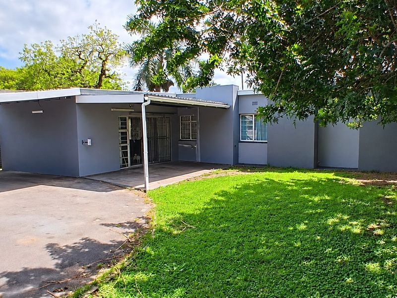 Rear view of the property featuring a covered patio area, paved driveway, and a well-maintained lawn with established trees.