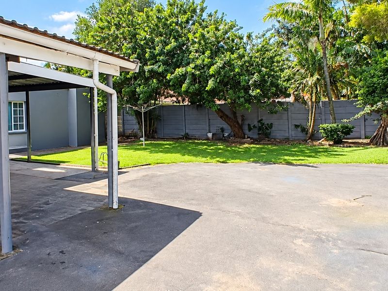 A spacious rear yard featuring a covered patio area, a large concrete driveway, and a well-maintained lawn enclosed by a boundary wall.