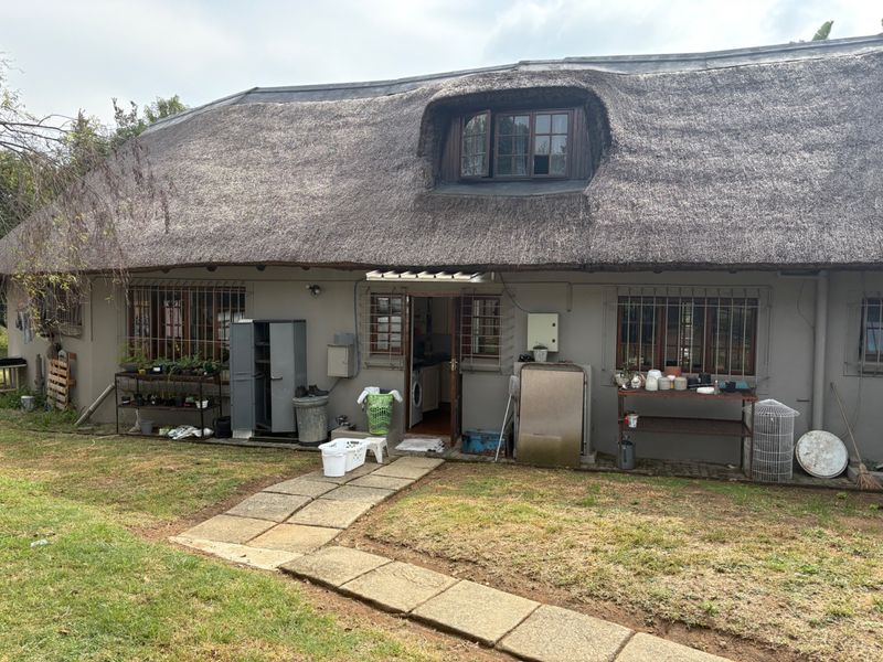 Rear view of a traditional thatched-roof cottage featuring a dormer window, security bars on windows, and a paved pathway leading to the entrance.