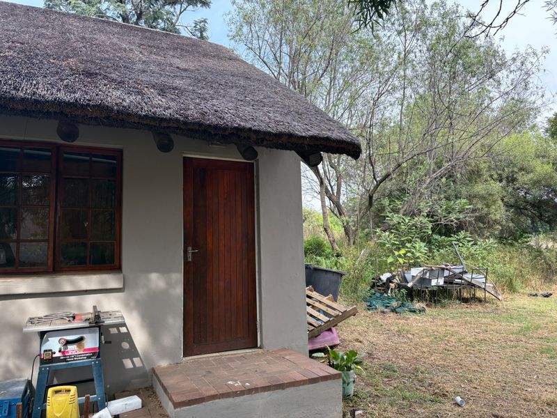 Rear view of a small holding structure featuring a traditional thatch roof, timber door, and brick entrance step set within a natural garden area.