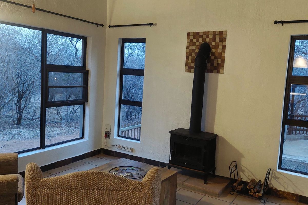 Living room featuring a freestanding fireplace, tiled flooring, and large aluminium windows offering natural light and views of the surrounding bushveld.
