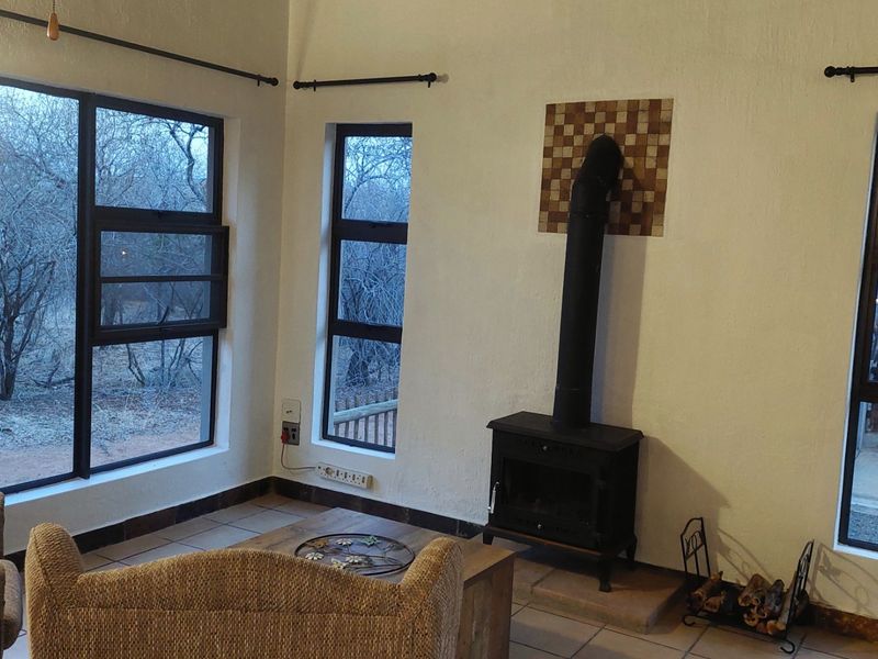 Living room featuring a freestanding fireplace, tiled flooring, and large aluminium windows offering natural light and views of the surrounding bushveld.