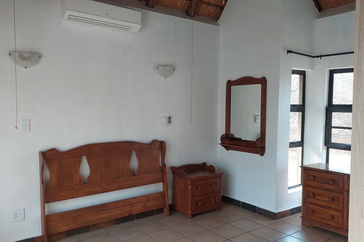 Bedroom featuring tiled flooring, exposed thatch ceiling, and a wall-mounted air conditioning unit with natural light from aluminium-framed windows.