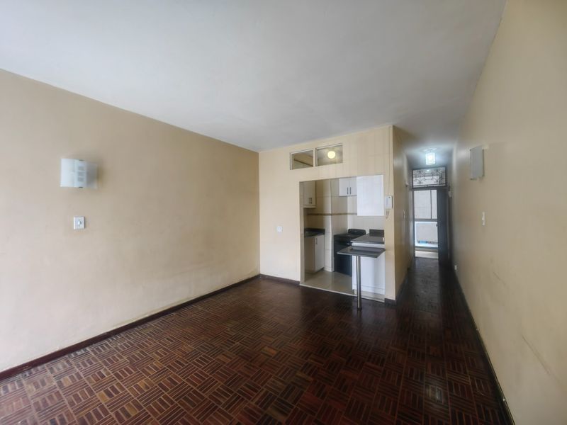 Open plan living area featuring parquet flooring and a compact kitchen with built-in cabinetry, leading to a hallway with a security gate.