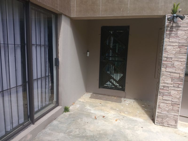 A covered patio area featuring a sliding glass door, security gate, and stone-clad pillar, requiring some maintenance to the concrete flooring and wall finishes.