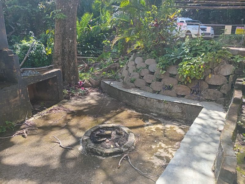 Outdoor entertainment area featuring a built-in braai, stone retaining wall, and a central concrete fire pit, requiring maintenance and landscaping.