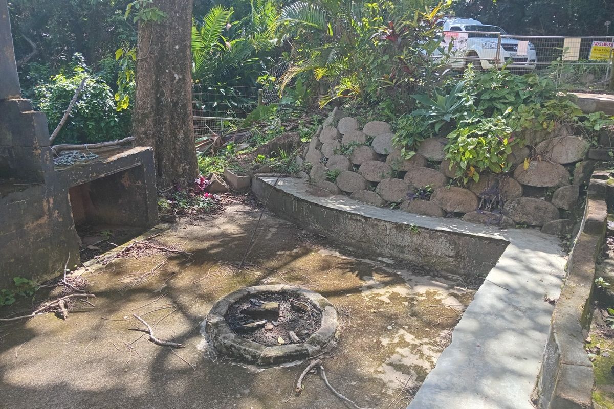 Outdoor entertainment area featuring a built-in braai, stone retaining wall, and a central concrete fire pit, requiring maintenance and landscaping.