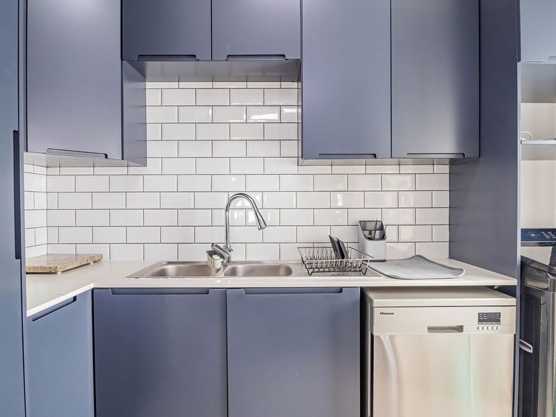 A modern, well-appointed scullery featuring dark blue cabinetry, white subway tile backsplash, stone countertops, and a stainless steel double sink with integrated dishwasher.