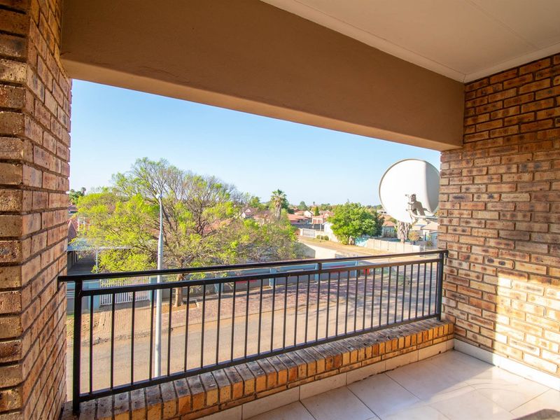 A covered balcony featuring exposed brick walls, a metal balustrade, and tiled flooring, offering an elevated view of the surrounding neighborhood.