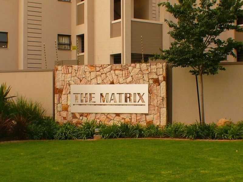 Front entrance view of The Matrix apartment complex featuring a stone-clad signage wall, manicured lawn, and perimeter electric fencing.