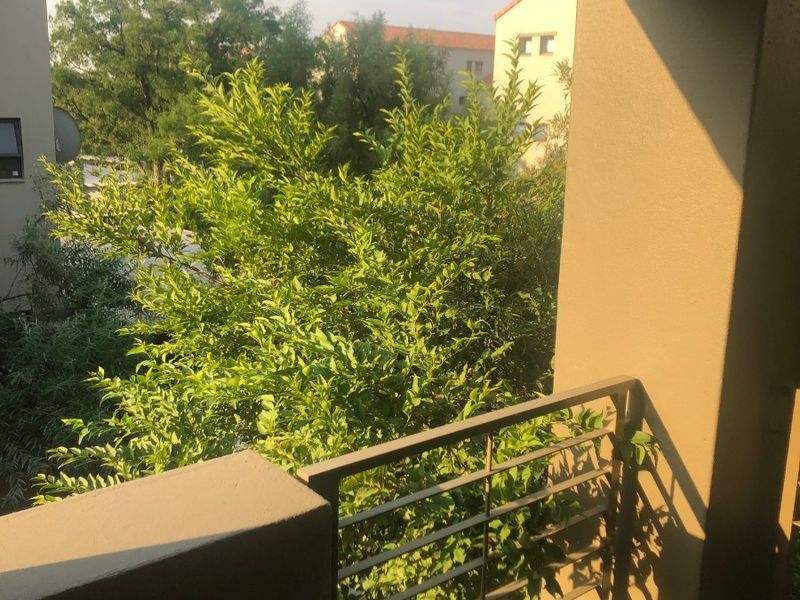 A covered balcony featuring a metal balustrade and concrete structural elements, offering a view of surrounding greenery and neighboring buildings.