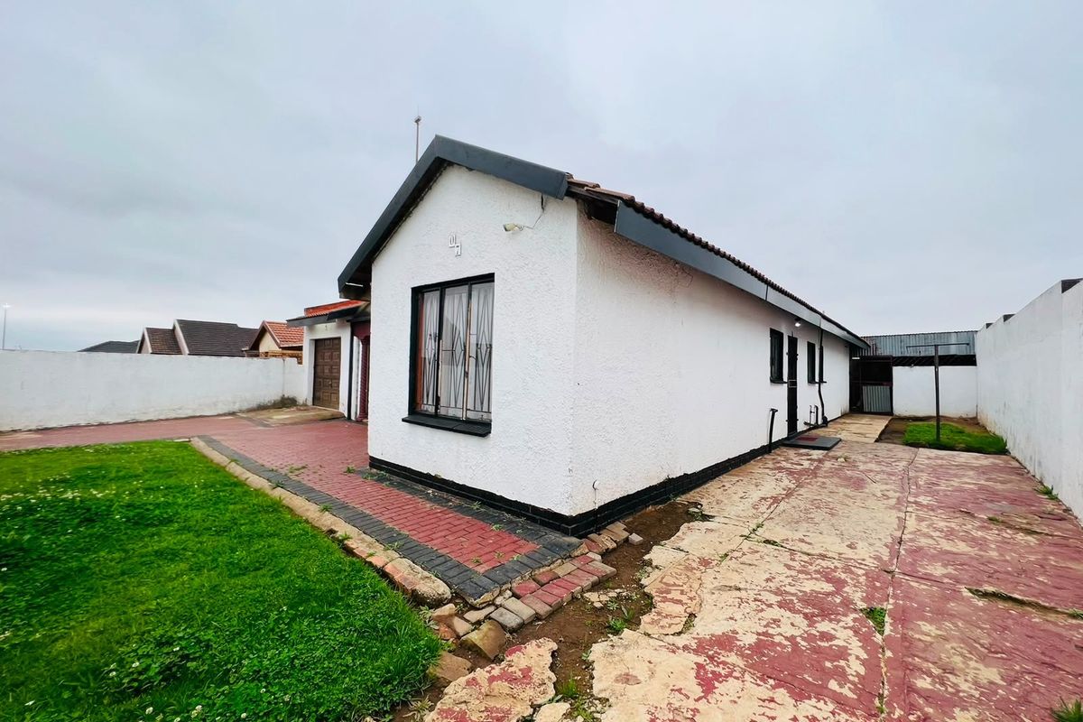 Rear view of the property featuring a white exterior, tiled roof, security bars on windows, paved driveway, and a small lawn area.