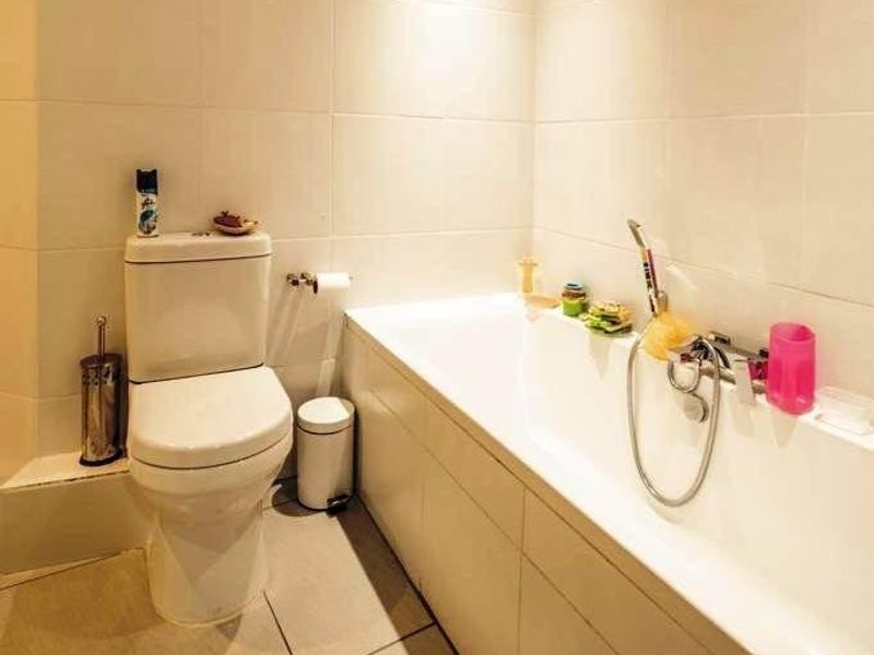 A clean, neutral-toned bathroom featuring a standard bathtub with a shower mixer and a modern toilet, finished with floor-to-ceiling wall tiles.