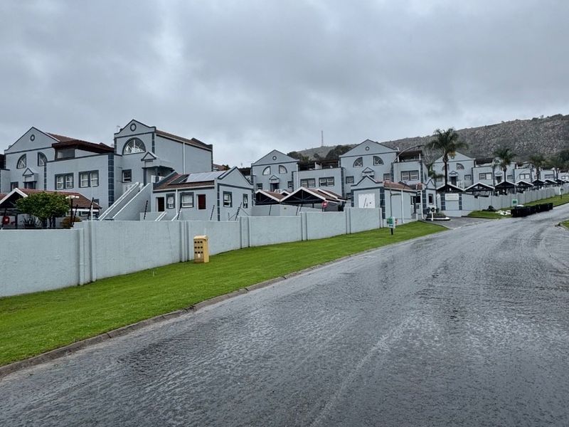 A street-level view of a townhouse complex featuring a paved road, manicured lawn, and perimeter wall with electric fencing.