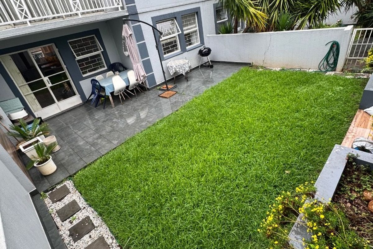 Rear exterior view of a townhouse featuring a tiled patio, a well-maintained lawn, and a balcony overlooking the private garden space.