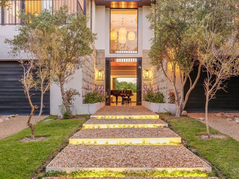 A striking modern entrance featuring stone cladding, illuminated gravel steps, and a grand double-volume glass facade leading into the home.