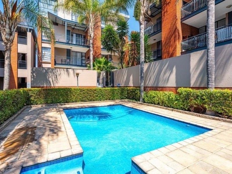 A well-maintained communal swimming pool area featuring paved decking, landscaped hedging, and surrounding apartment building architecture.