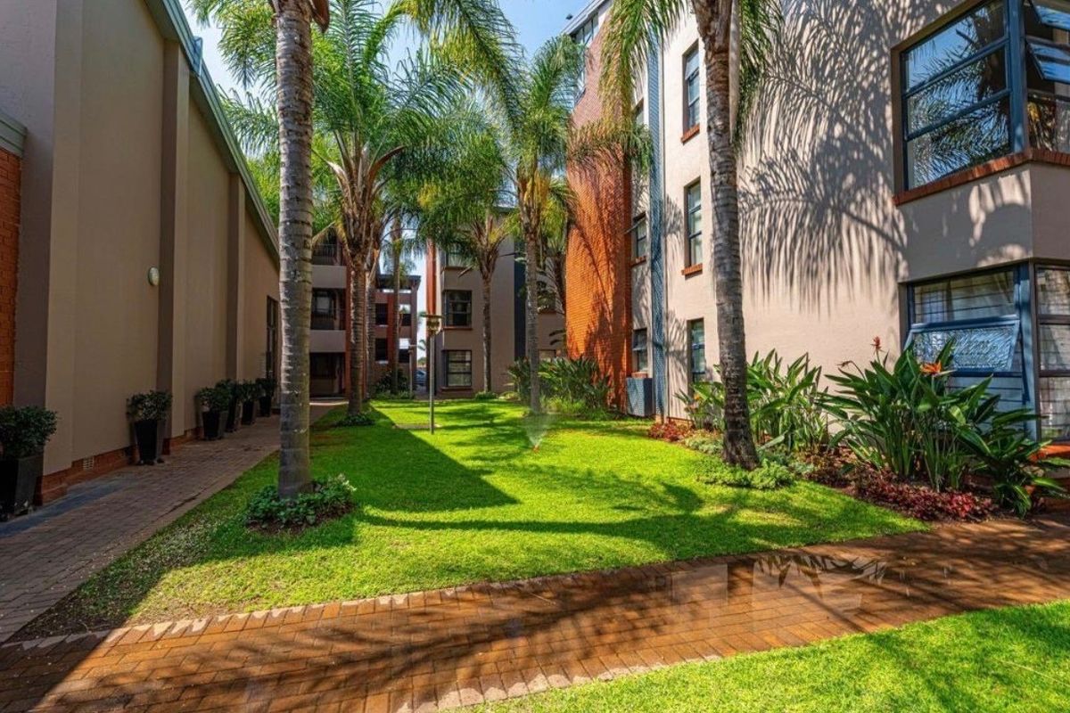 A well-maintained communal garden area between apartment blocks, featuring paved walkways, established palm trees, and manicured lawn.