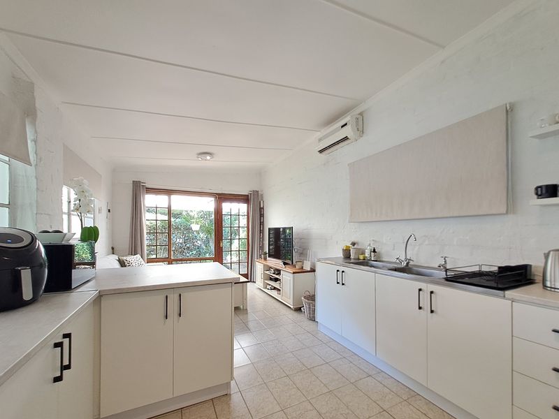 Open-plan kitchen and living area featuring white cabinetry, tiled floors, exposed brick walls, and direct access to the garden through french doors.