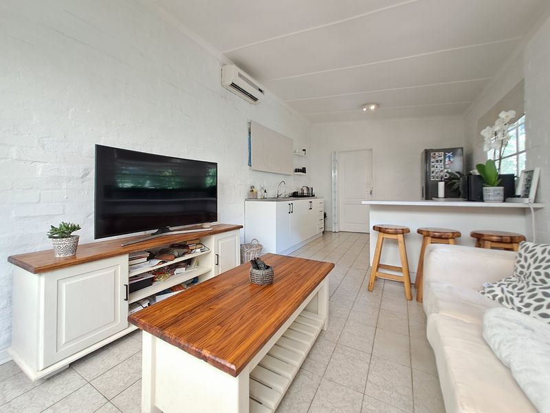 An open-plan living and kitchen area featuring tiled floors, white painted brick walls, a breakfast bar, and a wall-mounted air conditioning unit.