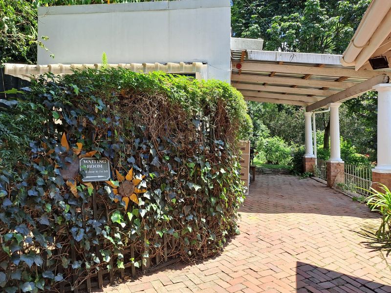 A covered outdoor patio area featuring brick paving and structural columns, surrounded by established garden greenery.