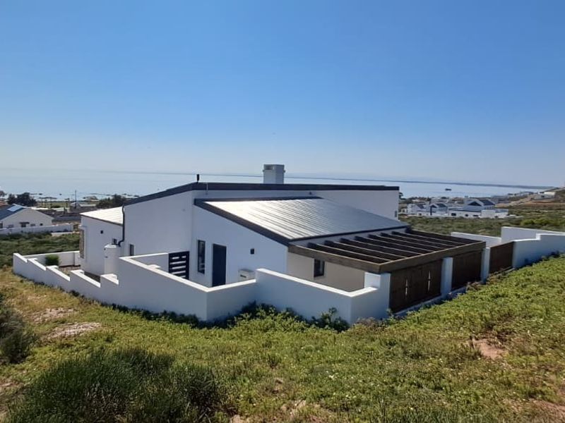 Elevated rear view of a modern coastal home featuring a metal roof, timber pergola, and surrounding boundary walls with scenic landscape views.