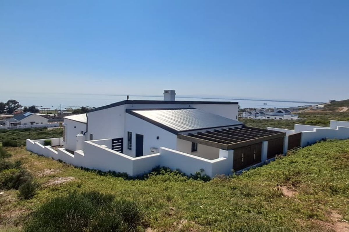Elevated rear view of a modern coastal home featuring a metal roof, timber pergola, and surrounding boundary walls with scenic landscape views.