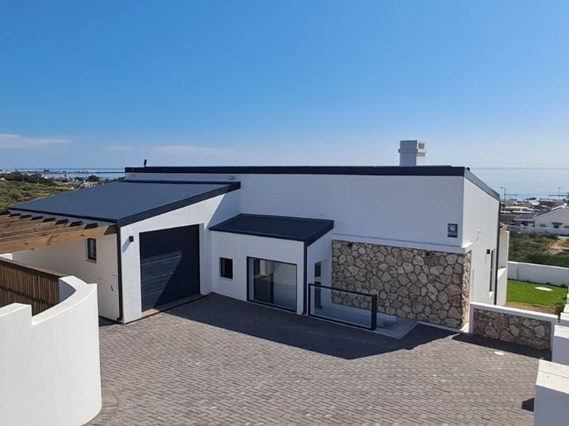 Modern coastal home featuring stone cladding, a paved driveway, integrated garage, and a timber pergola, set against a scenic landscape.