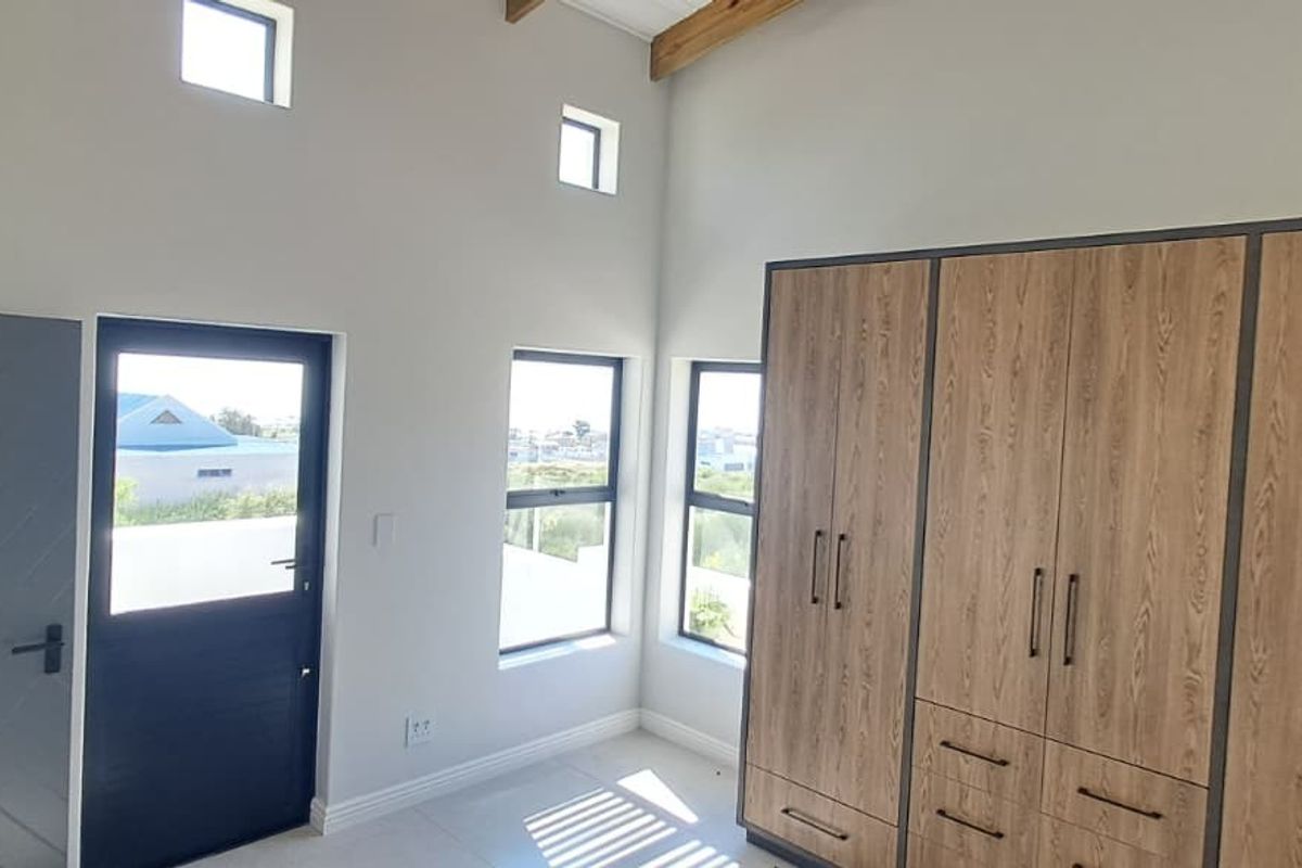 A bright, modern bedroom featuring high ceilings with exposed timber beams, tiled flooring, and extensive built-in wooden cabinetry.