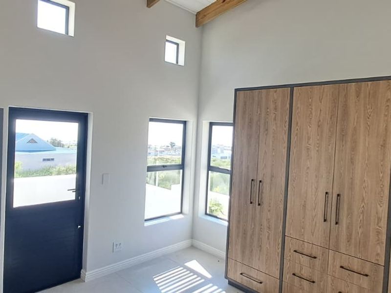 A bright, modern bedroom featuring high ceilings with exposed timber beams, tiled flooring, and extensive built-in wooden cabinetry.