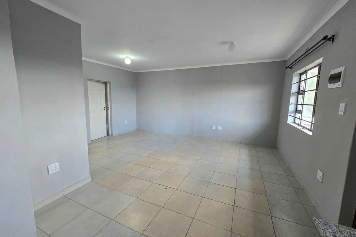 An empty, open-plan living area featuring neutral grey walls, tiled flooring, and security bars on the window.