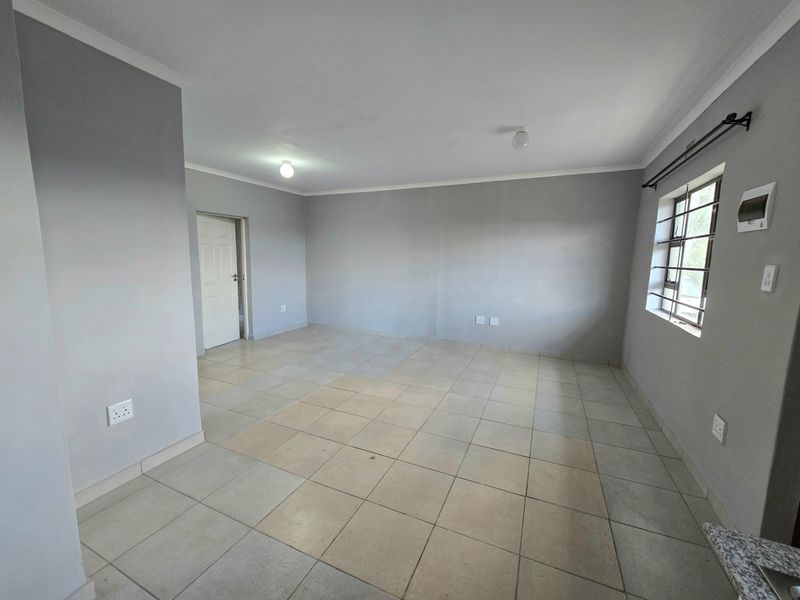 An empty, open-plan living area featuring neutral grey walls, tiled flooring, and security bars on the window.