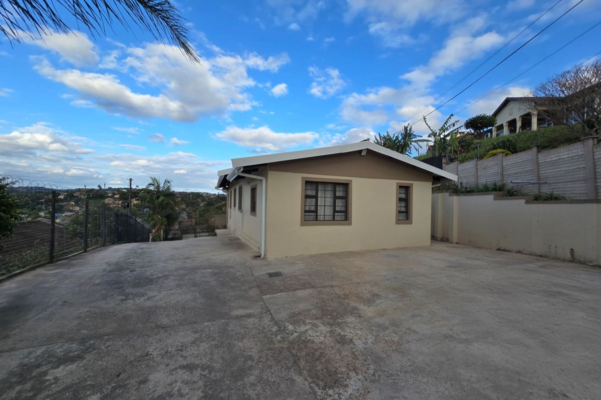 Exterior view of the property featuring a large concrete driveway, security fencing, and a retaining wall against a sloped landscape.