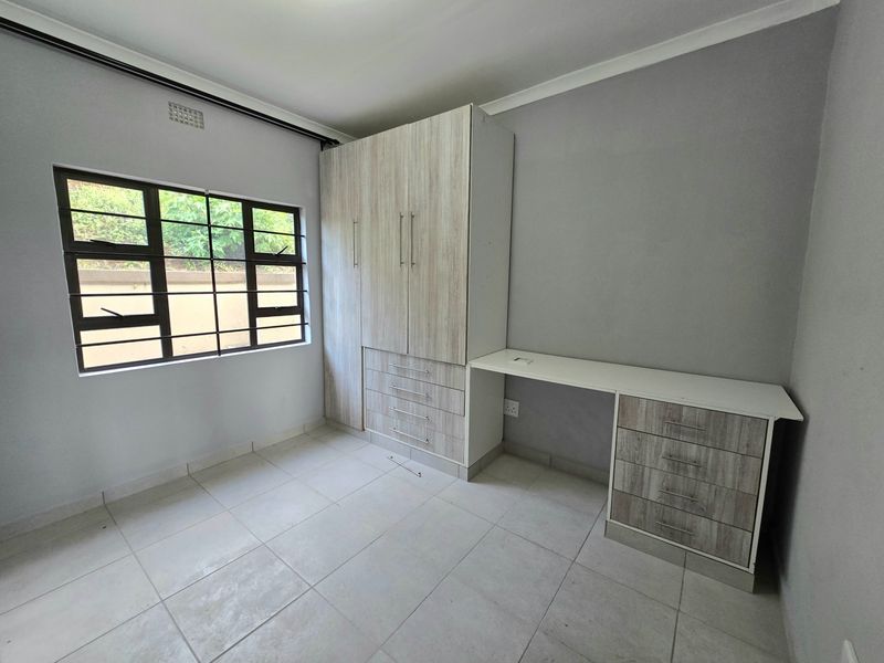 A bright, empty bedroom featuring neutral tiled flooring, built-in cupboards, and a matching integrated desk unit with ample storage.