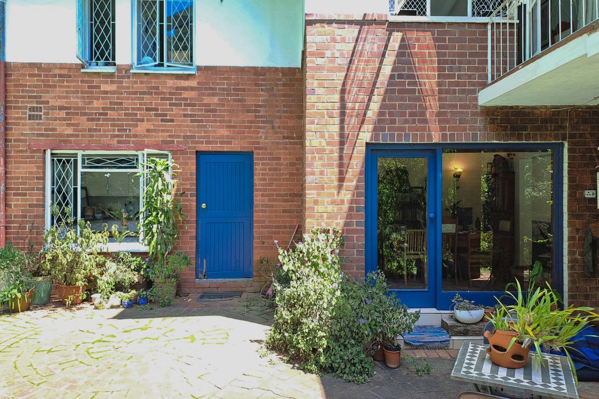 Rear exterior view of a brick cottage featuring a paved courtyard, blue entrance doors, and diamond-pattern security window bars.