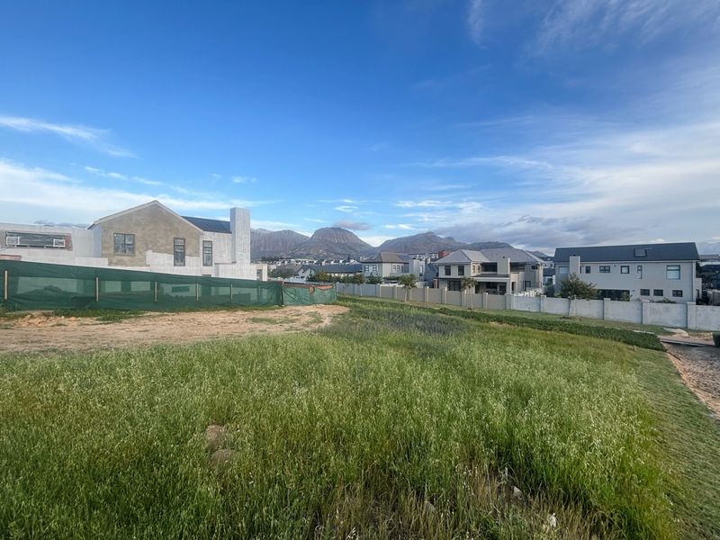 A vacant residential plot featuring natural grass cover, enclosed by perimeter walls, with surrounding residential development and mountain views in the background.