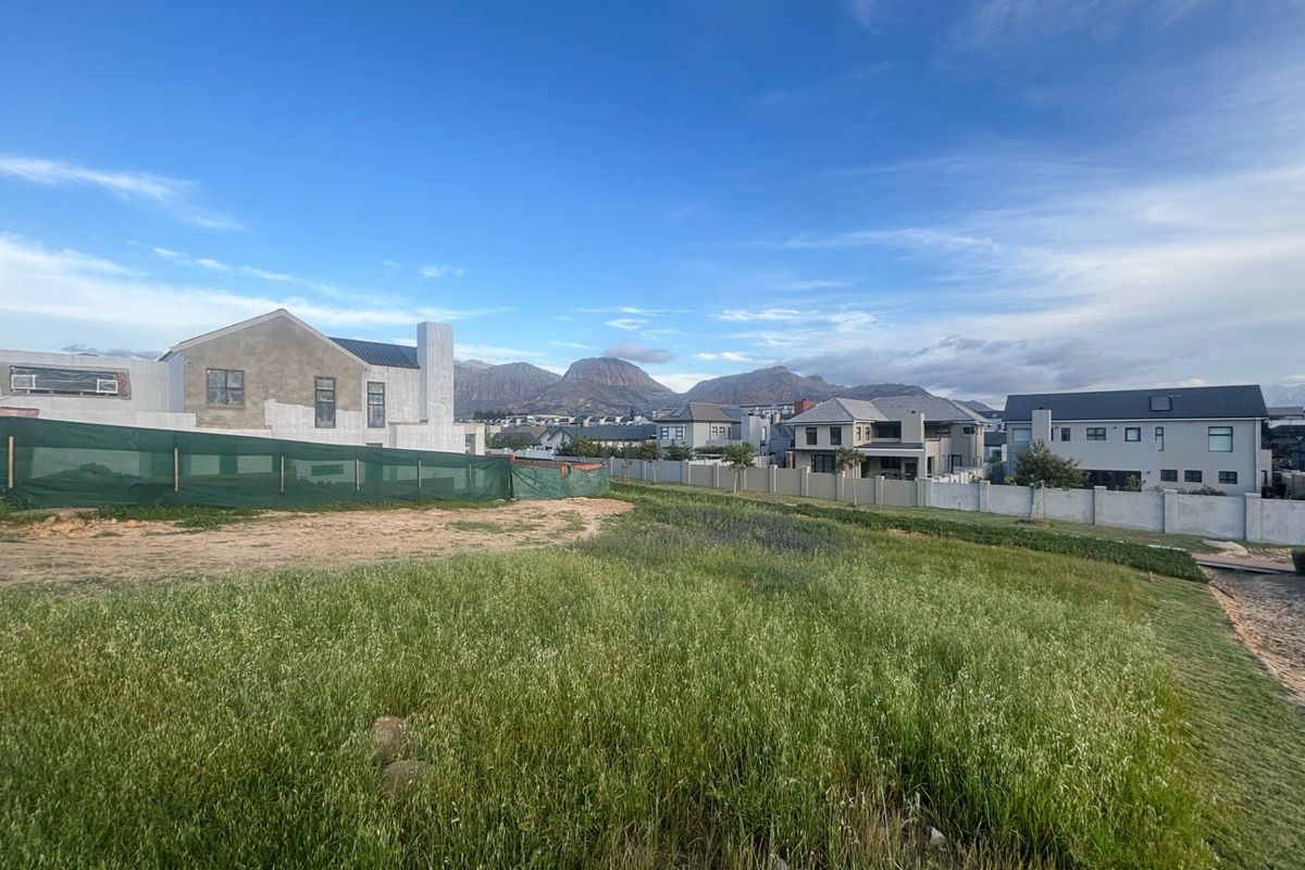 A vacant residential plot featuring natural grass cover, enclosed by perimeter walls, with surrounding residential development and mountain views in the background.