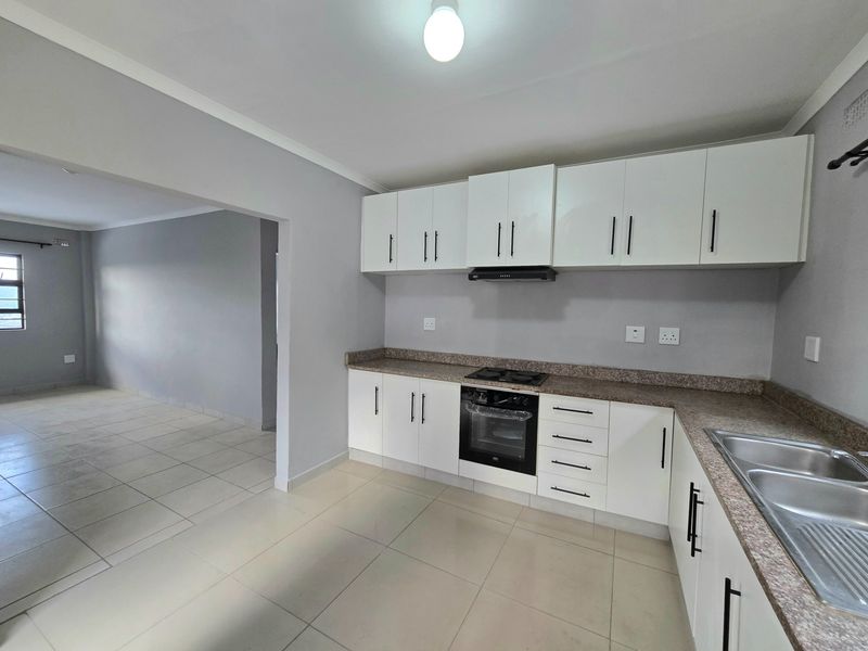 A clean, modern kitchen featuring white cabinetry, granite countertops, built-in oven, hob, and extractor fan, seamlessly flowing into an adjacent living area.