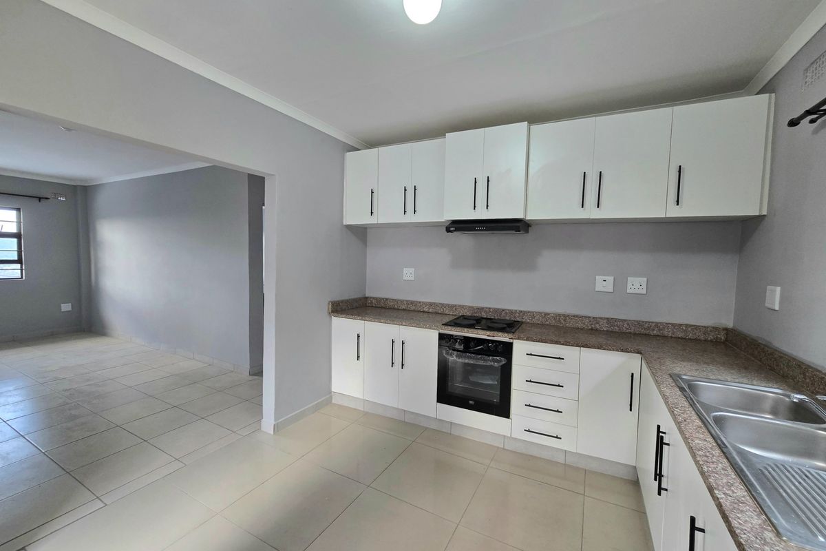 A clean, modern kitchen featuring white cabinetry, granite countertops, built-in oven, hob, and extractor fan, seamlessly flowing into an adjacent living area.