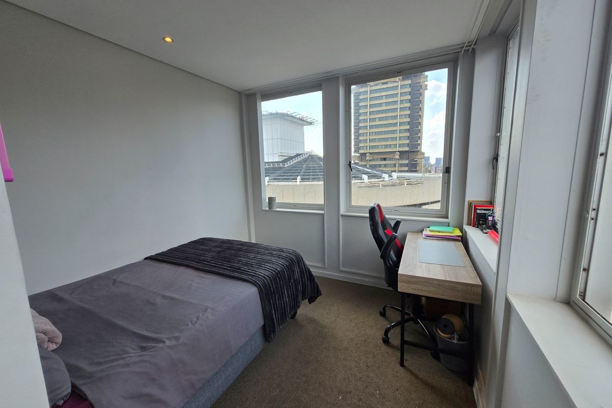 A compact bedroom featuring large windows with urban views, neutral wall finishes, and carpeted flooring.