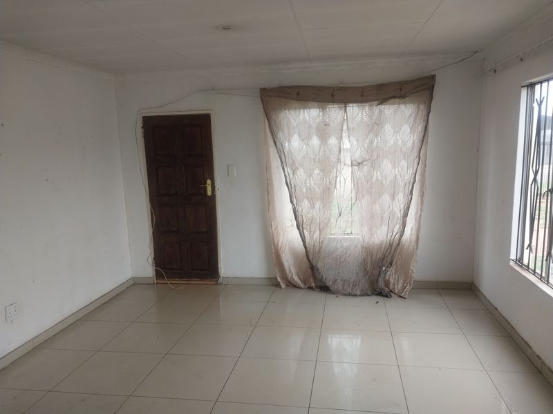 An empty living room featuring tiled flooring, a timber door, and window security bars, requiring cosmetic updates and ceiling repairs.