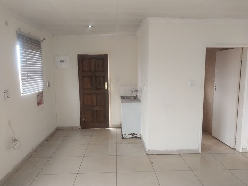 A basic, empty kitchen area featuring tiled flooring, a single basin sink with a damaged cabinet base, and a wooden exterior door.