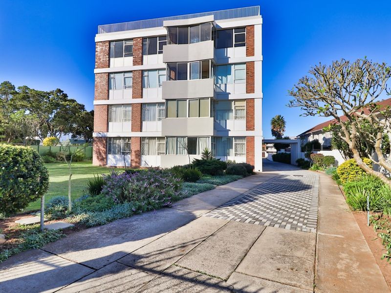Front view of a multi-story apartment building featuring a brick and concrete facade, paved driveway, and well-maintained surrounding garden area.