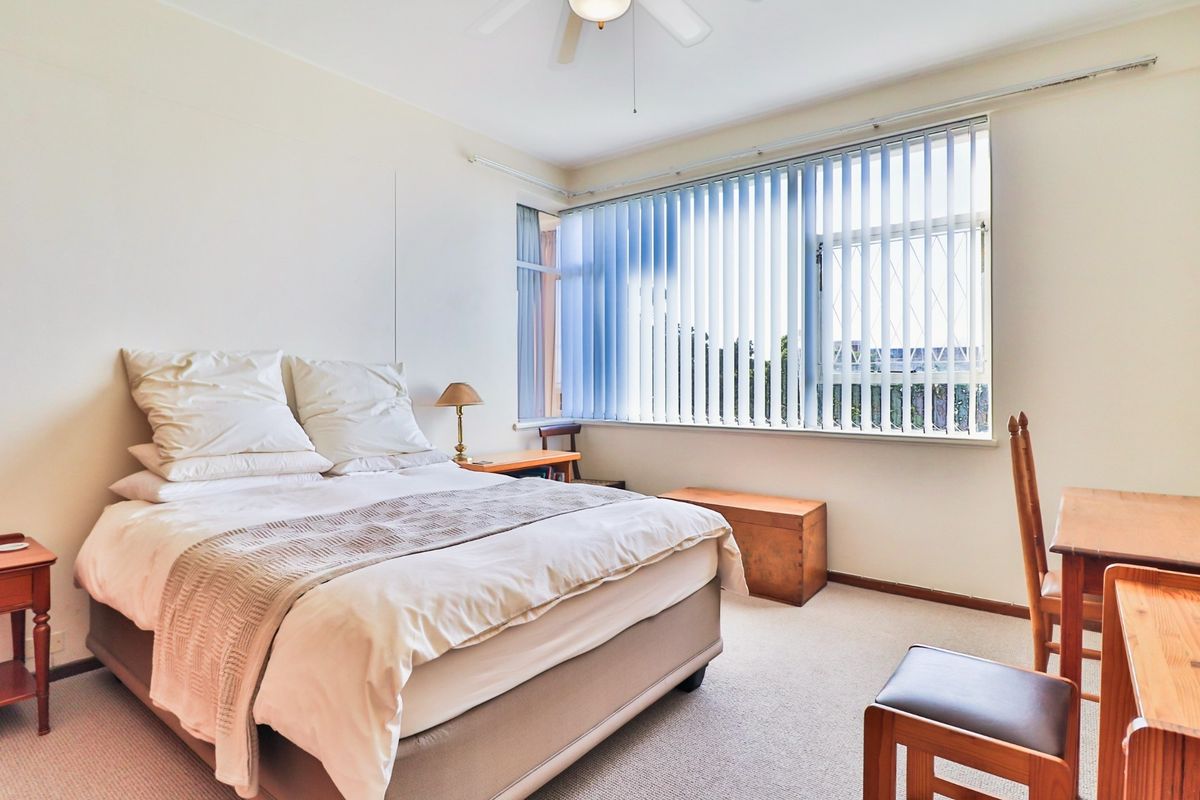 A well-lit bedroom featuring neutral carpet flooring, a ceiling fan, and large windows fitted with vertical blinds.