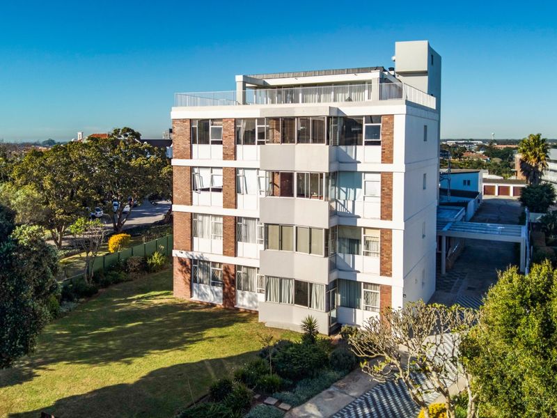 Aerial view of a multi-story apartment building featuring brick and plaster facade, balconies, a rooftop terrace, and surrounding landscaped garden areas.