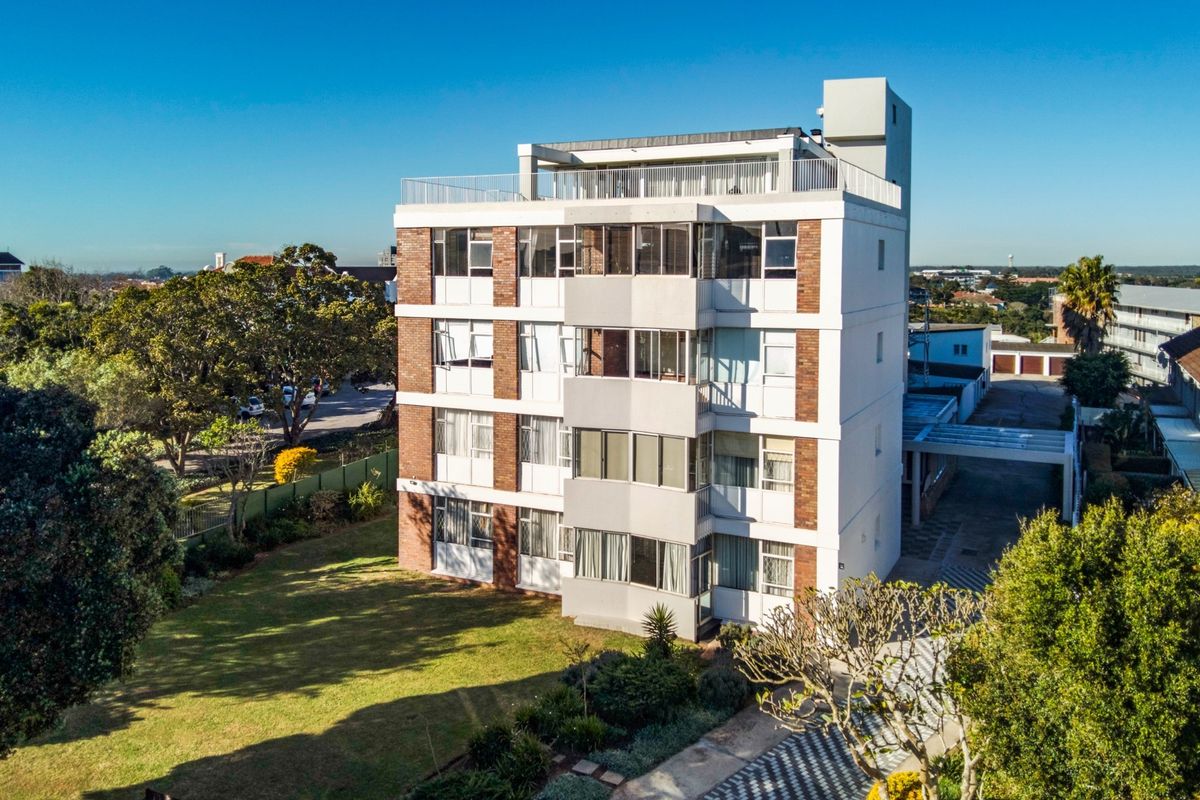 Aerial view of a multi-story apartment building featuring brick and plaster facade, balconies, a rooftop terrace, and surrounding landscaped garden areas.