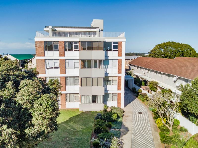Aerial view of a multi-story apartment building featuring a brick and concrete facade, paved driveway, and surrounding landscaped garden areas.