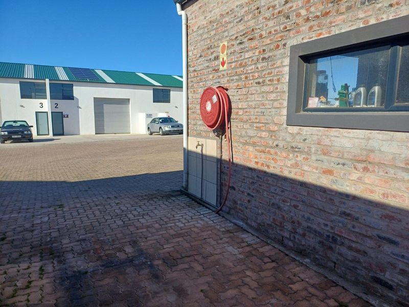 Exterior view of an industrial property featuring exposed brickwork, a fire hose reel, paved yard area, and adjacent industrial units with roller shutter doors.