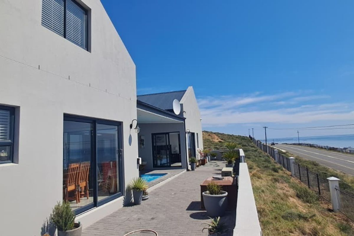 Rear exterior view of the property featuring a paved patio area, sliding glass doors, and a boundary wall overlooking the coastal road and ocean.